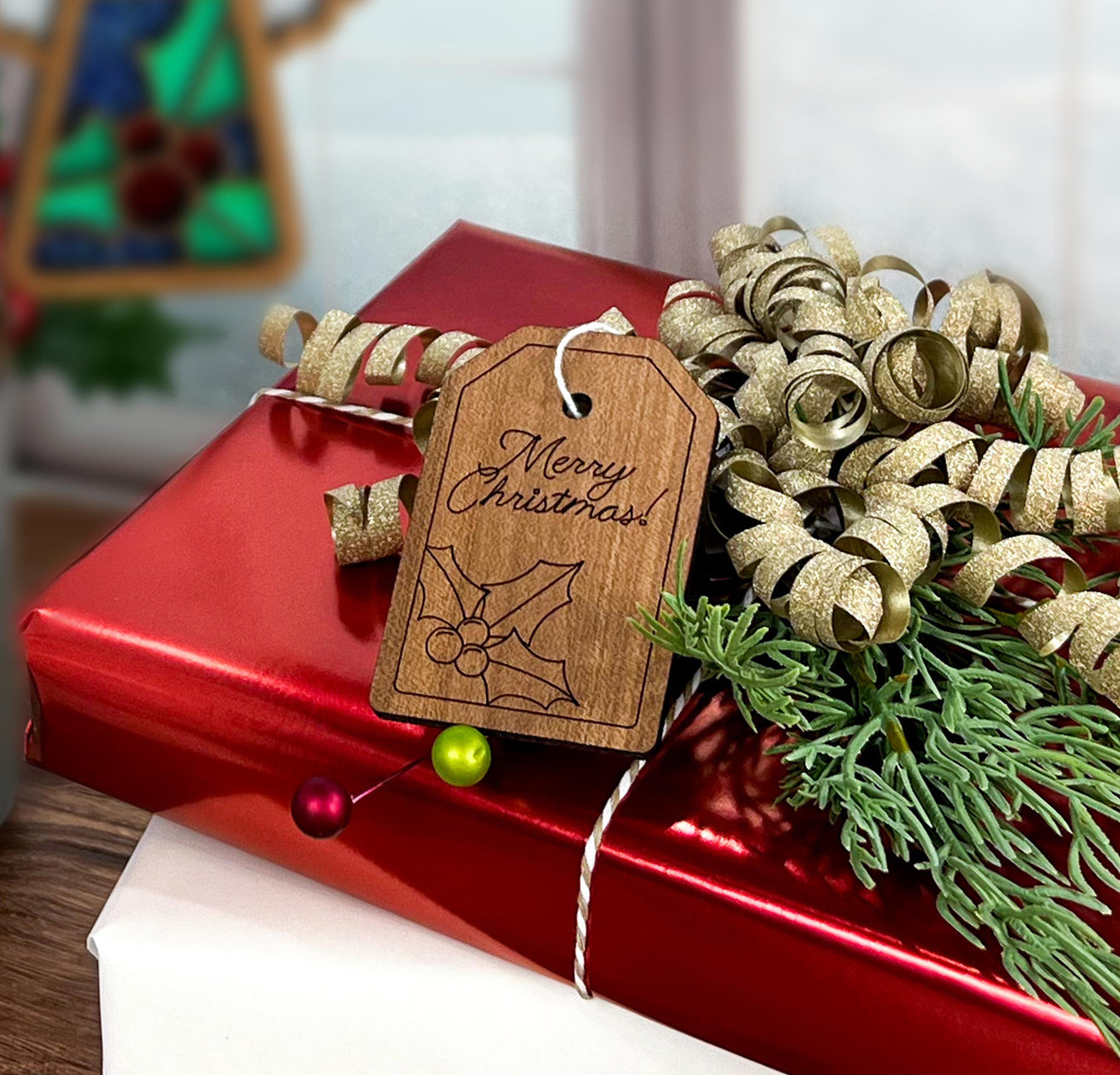 A festive Christmas present wrapped in red paper with a handcrafted Forged Flare® tag reading "Merry Christmas", accompanied by golden ribbons and greenery, ready to be adorned with Christmas angel ornaments.