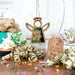 A Narcissus Ornament 3.5" Angel Figurine from Mother's Angels® by Forged Flare® is displayed among ribbon curls, a sprinkle-topped cupcake, a "Happy Birthday" tag, and scattered confetti on a wooden surface. Green gift-wrapped presents can be seen in the background.