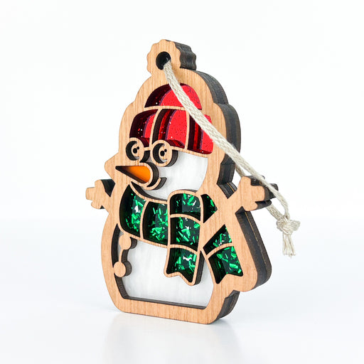 The Forged Flare® Christmas Snowman Ornament, 3.6", features a wooden snowman adorned with a red hat and green scarf. This festive decoration includes cut-out details highlighted by green glitter behind the scarf and a bright orange carrot nose. A string is attached for easy hanging, making it the perfect holiday decor.