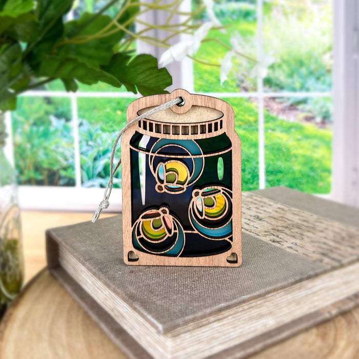 A Forged Flare® Fireflies Jar Ornament, 3.6", with colorful abstract designs sits on a stack of books. In the background, a window displays a lush green garden with plants, making it an ideal piece for summer home decor.