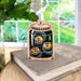 A Forged Flare® Fireflies Jar Ornament, 3.6", with colorful abstract designs sits on a stack of books. In the background, a window displays a lush green garden with plants, making it an ideal piece for summer home decor.