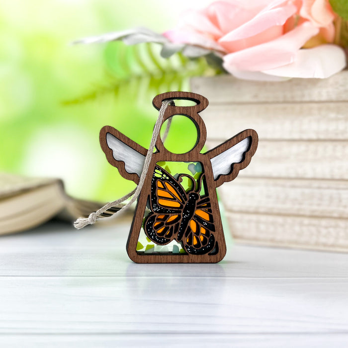 Displayed on a white surface is the Monarch Butterfly Ornament, a 3.5" angel figurine from Mother's Angels® by Forged Flare®. This small wooden angel features delicate white wings and a monarch butterfly ornament at its center. The background of blurred book pages and soft, pink flowers enhances the serene and peaceful atmosphere.