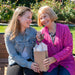 A young woman and an older woman sit on a bench outdoors, smiling happily. The older woman, adorned in a pink cardigan, holds up a small house-shaped keychain while the younger woman, wearing a denim jacket, holds a brown gift bag with tissue paper. They seem to be celebrating an occasion together, with the Forged Flare® Sunflower Ornament | 3.5" Angel Figurine | Mother's Angels® standing out as one of their unique gift ideas for women.