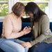 Two women sit closely on a blue couch, forehead to forehead, with eyes closed. One holds a small Daisy Ornament—a 3.5" Angel Figurine from the Mother's Angels® collection by Forged Flare®. They appear to be in a moment of heartfelt connection, offering mutual support and comfort as light streams in from the background windows.