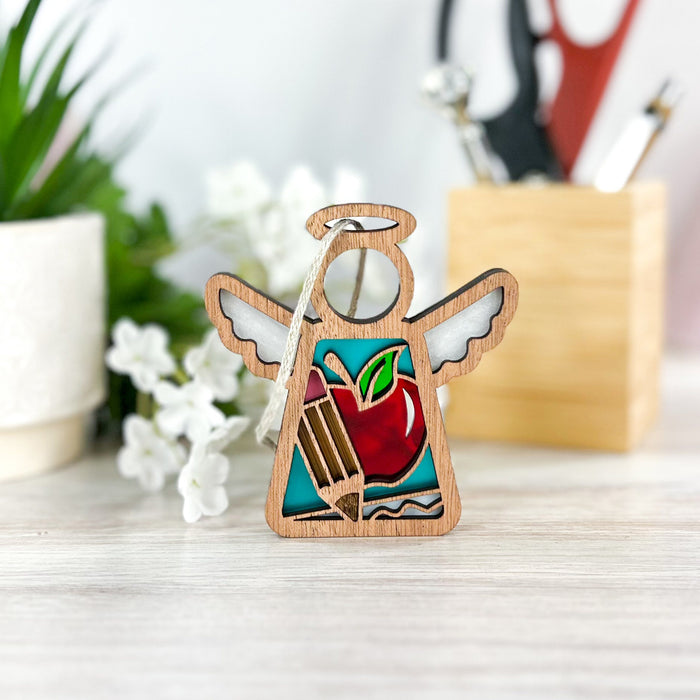 The Forged Flare® 3.5" Angel Figurine from the Mother's Angels® collection is displayed on a wooden surface. This beautiful wooden angel ornament, featuring cut-out wings, a red apple, and colored pencils, sits in front of a background with white flowers in a pot, green leaves, and blurred office supplies in a holder. Perfect as a Teacher Ornament or Christmas tree decoration.