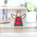 The Limited Edition Interlocking Hearts Ornament - Series 8, a 3.5" angel figurine from Mother's Angels® by Forged Flare®, features a glittery green background adorned with two pink heart-shaped embellishments. Perfect for Valentine's Day gifts, the wooden ornament is displayed on a wooden surface with a blurred background that includes a white potted plant and a pen.