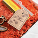 A close-up of the Forged Flare® 3" wooden gift tag featuring the message "For You. Just Because." surrounded by small flower drawings. The tag is attached with a red and white string to a package filled with orange shredded paper, with other items in the package partially visible.