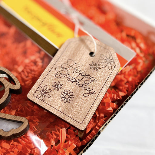 A close-up image of a Forged Flare® 3" wooden gift tag engraved with the message "Happy Birthday," adorned with intricate flower designs. The tag is attached with string and placed on a bed of shredded red paper, alongside other partially visible gift items.