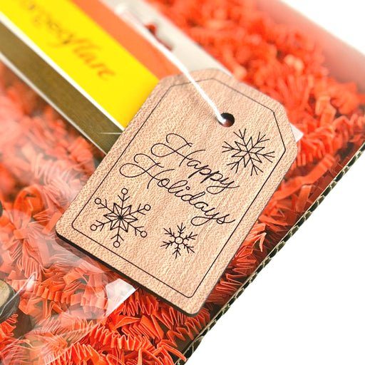A close-up of a holiday gift reveals an elegant Forged Flare® Gift Tag reading "Happy Holidays" in script. The 3-inch tag, adorned with snowflake illustrations, is nestled in red shredded filling inside a transparent box, signaling festive packaging and spreading a cheerful message.