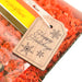 A close-up of a holiday gift reveals an elegant Forged Flare® Gift Tag reading "Happy Holidays" in script. The 3-inch tag, adorned with snowflake illustrations, is nestled in red shredded filling inside a transparent box, signaling festive packaging and spreading a cheerful message.
