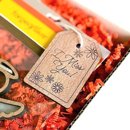 Close-up of a gift box filled with orange shredded paper. A brown Forged Flare® Gift Tag - "I Miss You!" 3" is attached to the box, featuring cursive text and decorated with drawings of flowers. Part of a colorful item with a yellow and orange gradient is visible inside the box, conveying a heartfelt message.