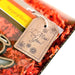 Close-up of a gift box filled with red shredded paper. A brown Forged Flare® Gift Tag - "I Miss You!" 3" is attached to the box, featuring cursive text and decorated with drawings of flowers. Part of a colorful item with a yellow and orange gradient is visible inside the box, conveying a heartfelt message.