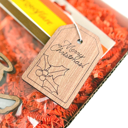 A close-up image of a package, adorned with a Forged Flare® Gift Tag engraved with "Merry Christmas!". The wooden tag, measuring 3 inches, features holly leaves and berries. The package is filled with orange shredded paper, adding a festive touch to the Merry Christmas message.