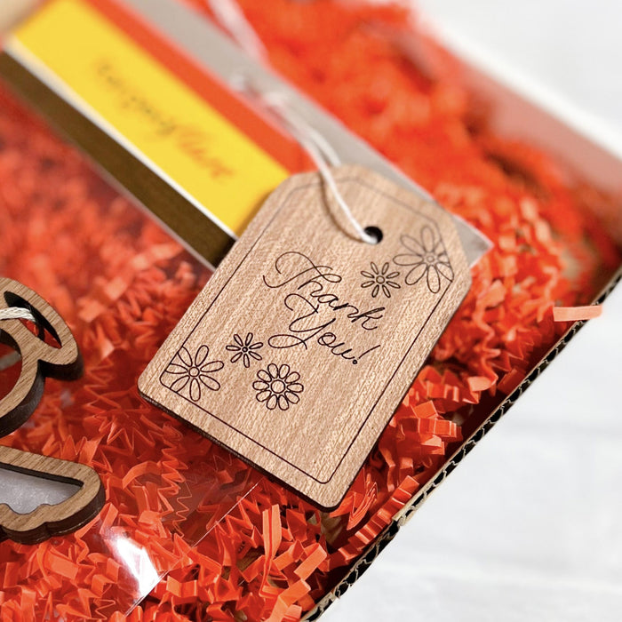 A "Thank You!" gift tag from Forged Flare®, featuring intricate floral designs etched onto sturdy wood and tied with white string, adorns a gift presented with orange crinkle paper and an angel ornament. This 3-inch tag beautifully conveys a sense of gratitude.