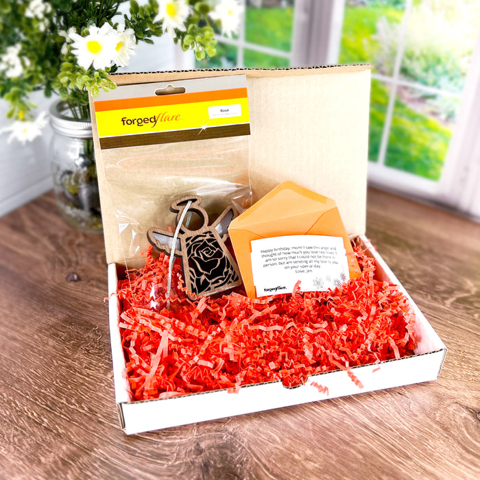 A Mother’s Angels® ornament displayed in an open box lined with orange crinkle paper on a wooden table. The set includes an ornament, a colored envelope, and a custom gift message card. A vase with white flowers and a window showing greenery are in the background.