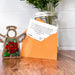 An orange envelope with a custom gift message card partially pulled out. The envelope leans against a jar of green foliage, and a small angel figurine featuring a rose design stands beside it on a wooden surface.