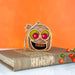 The Forged Flare® Halloween Monster - Mummy Ornament, standing at 3.5", is a small, colorful decoration placed on a brown book with a decorative ribbon bookmark. This cute mummy ornament features large green and pink eyes, and a wide smile. In the background, some decorative berries adorn the right side—making it a perfect Halloween gift or piece of spooky decor.