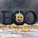 A Halloween decoration spells out "BOO" in black letters in the background, with a Forged Flare® Halloween Monster - Mummy Ornament, 3.5", featuring a mummy face, hanging from the second "O." Orange and white artificial berry branches are placed at the base, set against a dark, textured background. This makes for perfect spooky decor or a great Halloween gift.