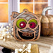 Introducing the Halloween Monster - Mummy Ornament by Forged Flare®, a 3.5" wooden decoration perfect for your spooky decor or as a unique Halloween gift. This charming ornament features a cartoonish mummy with pink and green eyes, a wide grin, and missing teeth. A braided linen string at the top makes it easy to hang, while its background showcases a window with a blurred pumpkin and white decorative berries.