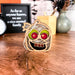 A 3.5" Halloween Monster Mummy Ornament from Forged Flare® rests on a wooden table, its eerie glow adding to the spooky atmosphere. Behind it, a framed sign declares, "As far as anyone knows, we are a nice normal family," accompanied by a small plant, a clock, and a box of assorted pens. This quirky piece makes an excellent Halloween gift for your unique decor needs.