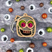 A Halloween Monster - Mummy Ornament, 3.5" from Forged Flare®, shaped like a pumpkin with a googly-eyed, smiling face featuring green and red eyes and glittery teeth, is placed on a textured gray surface surrounded by large googly eyes and small glittery balls in green, orange, and brown. It's an unexpected but delightful Halloween decoration idea.