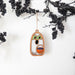 A whimsical Halloween ghost decoration from Forged Flare®, the 3.8" Trick-or-Treating Ghost Ornament, features colorful eyes and holds a small pumpkin as it hangs on a string amidst black berry branches against a white background. This glow-in-the-dark ornament adds an extra spooky touch to your seasonal decor.