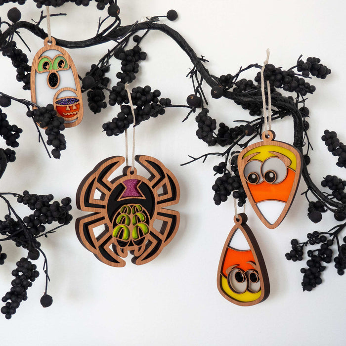 A decorative branch adorned with black berries hosts four colorful Halloween ornaments. These include two candy corns with eyes, a spider with green markings, and a 3.8-inch glow-in-the-dark ghost ornament by Forged Flare® featuring green eyes and a jack-o'-lantern mouth.