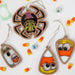 Four Halloween-themed wooden ornaments are laid out on a white background surrounded by candy corn and plastic googly eyes. Among them is a spider with a patterned back, three candy corn pieces with various facial expressions, and the Halloween Trick-or-Treating Ghost Ornament from Forged Flare®, which glows in the dark.