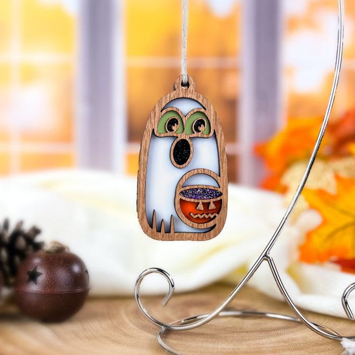 The Halloween Trick-or-Treating Ghost Ornament from Forged Flare® hangs on a silver stand, featuring green eyes and a pumpkin face. This 3.8" wooden ghost ornament glows in the dark, adding an eerie charm. It is set against a backdrop of autumn decor with pinecones and orange leaves, complemented by a blurred window view.