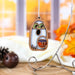 The Halloween Trick-or-Treating Ghost Ornament from Forged Flare® hangs on a silver stand, featuring green eyes and a pumpkin face. This 3.8" wooden ghost ornament glows in the dark, adding an eerie charm. It is set against a backdrop of autumn decor with pinecones and orange leaves, complemented by a blurred window view.