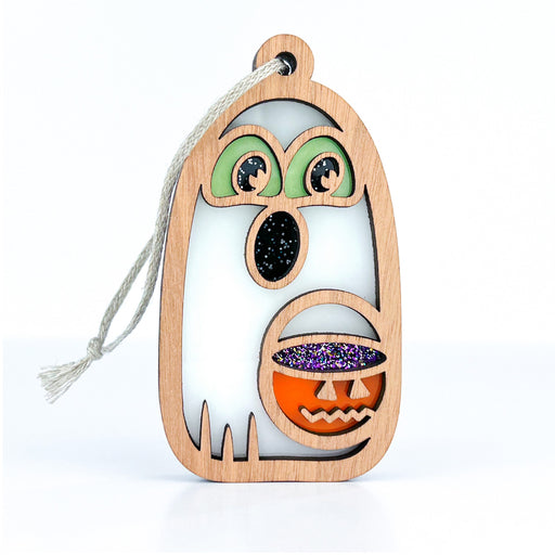 The Halloween Trick-or-Treating Ghost Ornament by Forged Flare®, measuring 3.8 inches, is a wooden decoration shaped like a ghost with cut-out eyes featuring green and black accents. It holds a small jack-o'-lantern bucket adorned with purple and orange glitter. A string loop is attached at the top for hanging, making this glow-in-the-dark ornament perfect for spooky nights.