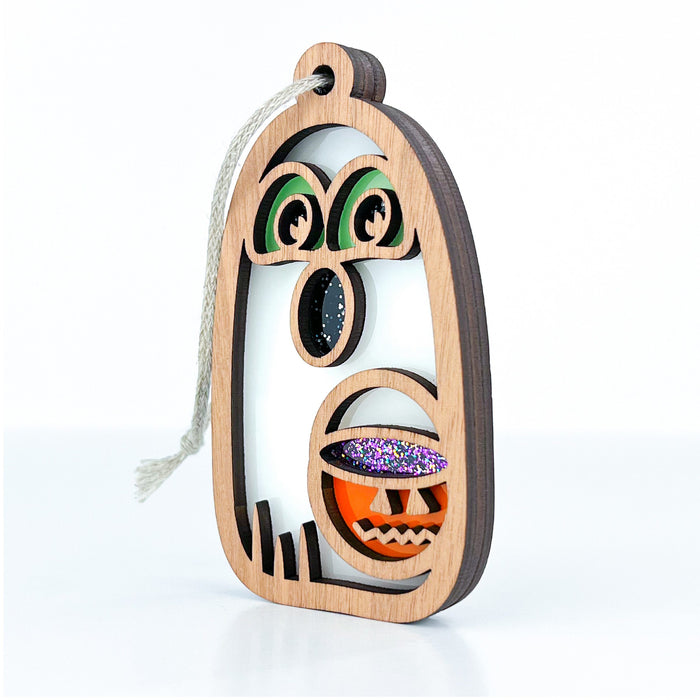 The Forged Flare® Halloween Trick-or-Treating Ghost Ornament, 3.8", is a wooden decoration shaped like a ghost or monster featuring green eyes and a mouth filled with colorful, glittery shapes. It includes a braided linen string at the top for easy hanging, making it the perfect glow-in-the-dark Halloween ghost decoration.