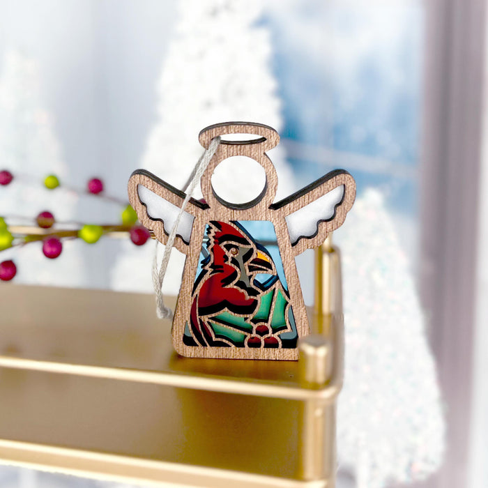 A Winter Cardinal Ornament from Forged Flare®'s Mother's Angels® collection stands on a golden shelf. The 3.5" wooden angel figurine with wings and a circular halo features a colorful stained-glass-style depiction of a red cardinal perched on a branch, making it an ideal gift for bird lovers. The background includes blurred holiday decorations and white Christmas trees.