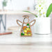A Forged Flare® Mother's Angels® 3.5" Angel Figurine stands on a light-colored table, making a perfect birthday gift. The angel features a stained-glass section showcasing yellow daffodil flowers. In the background, there's a blurred white flower pot with green leaves and small white blooms, alongside a pen on a shelf.