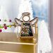 The Narcissus Ornament by Forged Flare®, a 3.5" angel figurine from the Mother's Angels® collection, features a delicate design and stands on a golden surface. In the background, blurred white Christmas trees with a snowy appearance and a string of red and green beads create a festive scene. The angel has white wings and a simple halo, embodying the peaceful beauty of December's birth flowers.