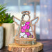 Introducing the Primrose Ornament | 3.5" Angel Figurine by Mother's Angels®. This enchanting ornament features a decorative wooden angel with wings and a body adorned with pink flowers, beautifully accented with yellow centers. It is elegantly displayed on a wooden slice stand against a blurred backdrop that includes a plant. Brought to you by Forged Flare®, it makes for the perfect last-minute Mother's Day gift for mom.