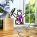 The Primrose Ornament, a 3.5" angel figurine from the Mother's Angels® collection by Forged Flare®, is adorned with intricate floral cutouts and purple accents. It hangs gracefully from a silver stand, set against a backdrop of blurred office supplies, purple flowers, and a bright window with blurred greenery. This delicate piece is an ideal birthday gift for women.