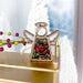 A Red Rose Ornament | 3.5" Angel Figurine from the Mother's Angels® collection by Forged Flare® features a wooden angel with vibrant stained glass wings and a red floral design, resembling a birth flower, resting on a gold surface. In the backdrop, softly blurred white Christmas trees and festive decorations adorned with green and red berries add to the holiday charm.