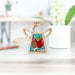 The Teacher Ornament by Forged Flare® is a charming 3.5" Mother's Angels® figurine, featuring a small wooden angel with cutout wings, a halo, and body adorned with colorful inlays of an apple and pencils. Positioned on a light-colored surface with a blurred background that includes a white pot of flowers and a pencil on the desk, it makes the perfect teacher Christmas ornament.