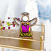 A 3.5-inch Tulip Ornament angel figurine from the Forged Flare® Mother's Angels® collection stands on a golden surface, featuring stained glass wings and a halo. Adorned with a purple tulip design, this enchanting Christmas tree ornament is surrounded by blurred white Christmas trees and festive red berries in the background.