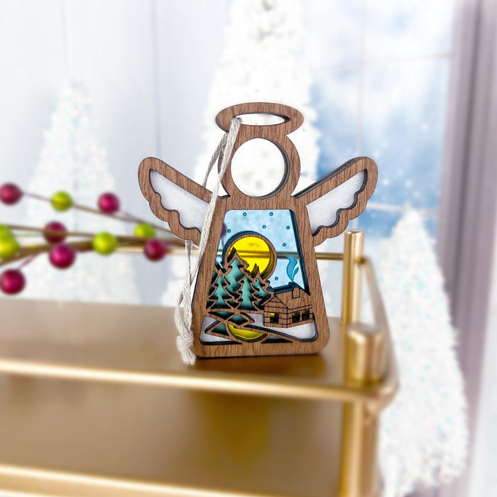 The Warm Welcome Ornament from Forged Flare®'s Mother's Angels® collection is a 3.5" angel-shaped wooden figurine. It features a intricate cut-out scene of a snowy log cabin nestled in a forest under the glow of a yellow moon, complete with detailed trees, creating a serene and peaceful setting. Displayed on a gold surface with blurred festive decorations and snowy trees in the background, this ornament brings an enchanting touch to any holiday display.