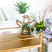 A Water Lily Ornament from Forged Flare's Mother's Angels® collection, a 3.5" angel figurine with white wings and a stained glass center showcasing a colorful water lily design, sits on a gold shelf. In the background, there is a potted green plant, some colorful birth flowers in a glass jar, and neatly arranged books on white shelves.