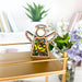 A Yellow Rose Ornament 3.5" Angel Figurine from Forged Flare's Mother's Angels® collection rests on a gold-colored shelf. In the background, there are books, white and purple flowers in a jar, and a white wall, creating a cozy, decorative setting.
