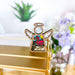 A Forged Flare® Mother's Angels® Flamingo Beach Ornament, a 3.5" wooden angel figurine with white wings, intricately painted with a vibrant pink flamingo in the center, resting on a gold surface amidst blurred greenery and purple flowers in the background.