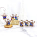 The image features six 3.5" Mother's Angels® wooden figurines from the Christmas Nativity Set Bundle by Forged Flare®, showcasing colorful and intricate designs on a marble surface. Three of the angels are arranged on a small wooden platform, two rest on a straw-like platform, and one stands alone. In the background, white Christmas trees add to the festive ambiance, making these nativity ornaments an ideal gift for the holiday season.