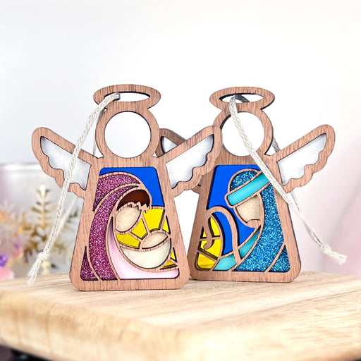 Two Mother's Angels® figurines from the Christmas Nativity 2-Piece Bundle by Forged Flare® rest on a wooden surface. Each 3.5" wooden angel ornament features colorful designs and cradles the Holy Family—Mary, Joseph, and baby Jesus—with one adorned in pink accents and the other in blue. These festive ornaments are equipped with strings for hanging.