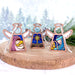 The Christmas Nativity 3-Piece Bundle from Forged Flare®, featuring star, Mary, and Joseph angel figurines from the Mother's Angels® collection, rests on a wooden surface. Snowy trees appear in the background, creating a festive and serene atmosphere. Each 3.5" angel has outstretched wings and a halo.