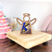 A wooden figurine shaped like an angel, complete with cut-out wings and a hanging loop, rests on a small wooden platform. This 3.5" nativity piece, known as the Christmas Nativity Purple Wise Man from Forged Flare's Mother's Angels® collection, showcases vibrant inlaid designs featuring a hat and potion bottle. In the background is a decorative pink Christmas tree amid various blurred objects. Ideal for enhancing your nativity scene or as a thoughtful gift for the season.