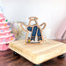 The Winter Snow Angel Ornament, a 3.5" angel figurine from the Mother's Angels® collection by Forged Flare®, is displayed on a small wooden stand. This wooden Christmas decoration features blue glitter accents and small gold stars. To the left, a pink, bejeweled Christmas tree and a festive mug are blurred in the background, creating a cozy holiday atmosphere. It makes for one of the perfect gifts for mom.