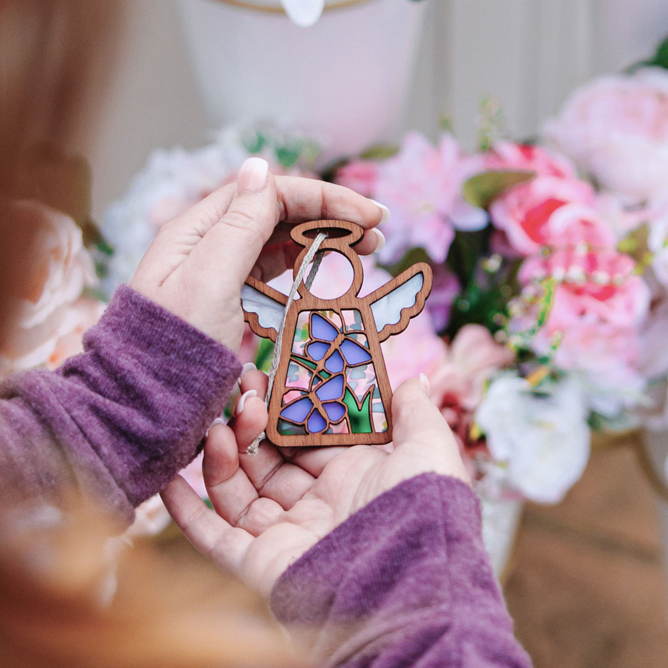 A person with reddish-brown hair and wearing a mauve-colored sweater holds the Playful Purple Butterflies Ornament, a 3.5" angel figurine from the Mother's Angels® collection by Forged Flare®. The delicate stained glass–style angel is intricately adorned with floral designs in purple and green, complemented by delicate butterfly decorations. In the background, there are pink and white flowers.